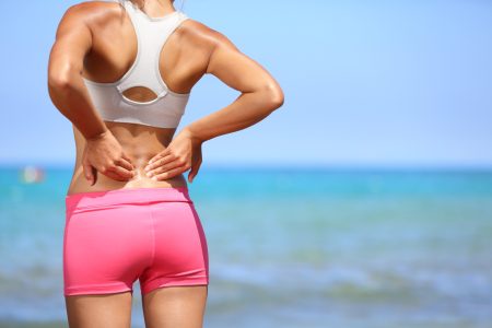 Athletic woman in pink sportswear standing at the seaside rubbing the muscles of her lower back, cropped torso portrait.